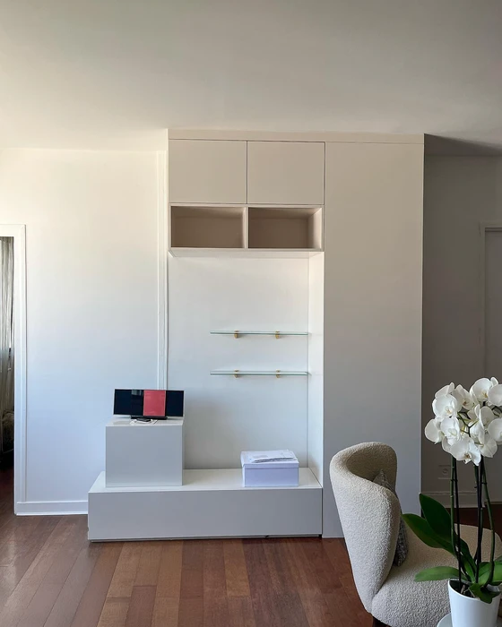 Salon minimaliste avec meuble mural sur mesure blanc mat, comprenant placards supérieurs à portes affleurantes, niches ouvertes, étagères en verre trempé avec supports dorés
