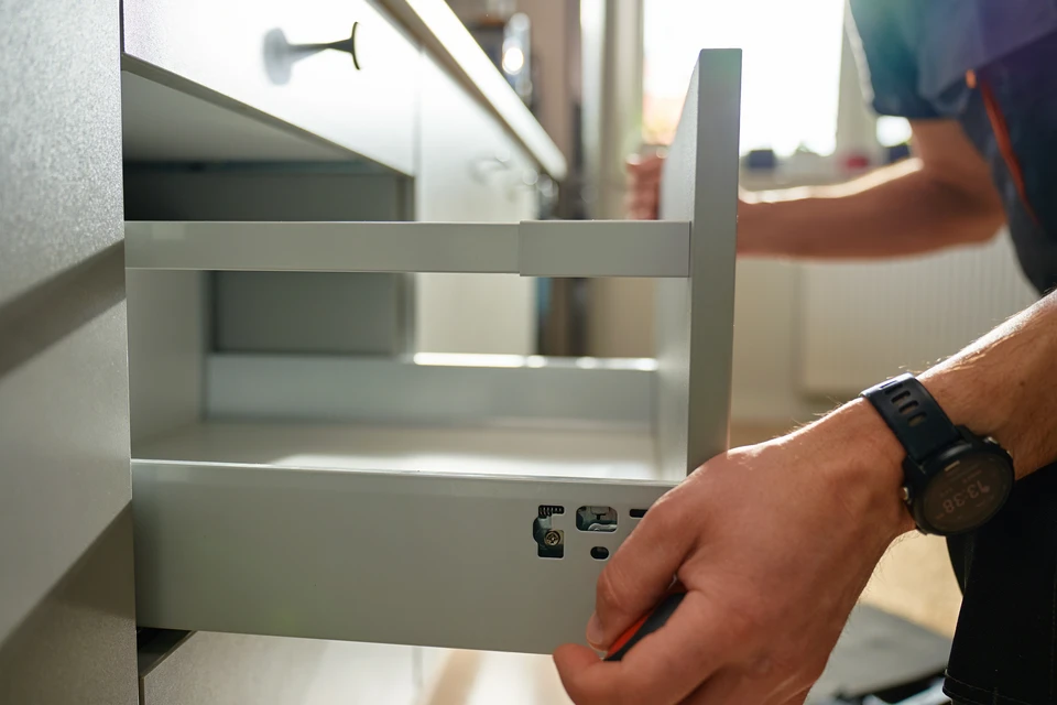 Photographe réaliste d'un artisan menuisier en tenue de travail, concentré sur son établi ou lors de l'installation d'une cuisine haut de gamme, avec des matériaux nobles visibles (bois, quincaillerie de qualité). La scène doit refléter la précision, l'exigence et le professionnalisme. On peut voir des détails de menuiserie fine, un environnement intérieur, et une attitude de contrôle qualité méticuleux. L'image doit transmettre l'excellence et l'engagement personnel du professionnel envers son ouvrage.