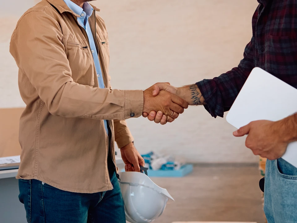 Photographie réaliste d'une artisan/designer en menuiserie accueillant un client dans un showroom ou atelier professionnel, tous deux regardant ensemble des plans de mobilier ou de cuisine sur une table lumineuse ou écran, avec des exemples de travaux finalisés visibles en arrière-plan dans un cadre haut de gamme et épuré. L'image doit transmettre confiance, professionnalisme et accompagnement personnalisé du projet.